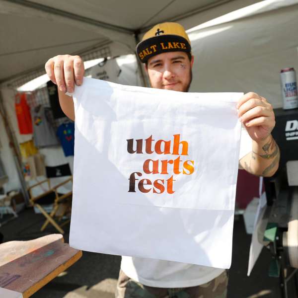person in salt lake hat holding up a utah arts fest t-shirt